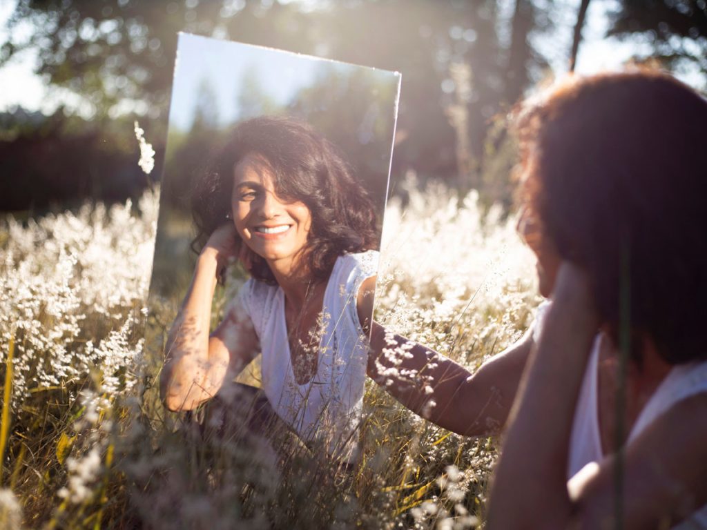 Photo of a woman in a field holding a mirror with her reflection. This represents how individual therapy can help you find true love with another person and yourself.