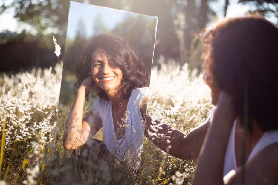 Photo of a woman in a field holding a mirror with her reflection. This represents how individual therapy can help you find true love with another person and yourself.