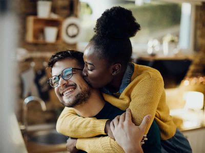 Photo of a multiracial couple hugging while the woman kisses her partner in the cheek in a tender moment. This represents how Couples Workshops And Intensive Couples Therapy Can Help you and your partner rediscover intimacy and connection.